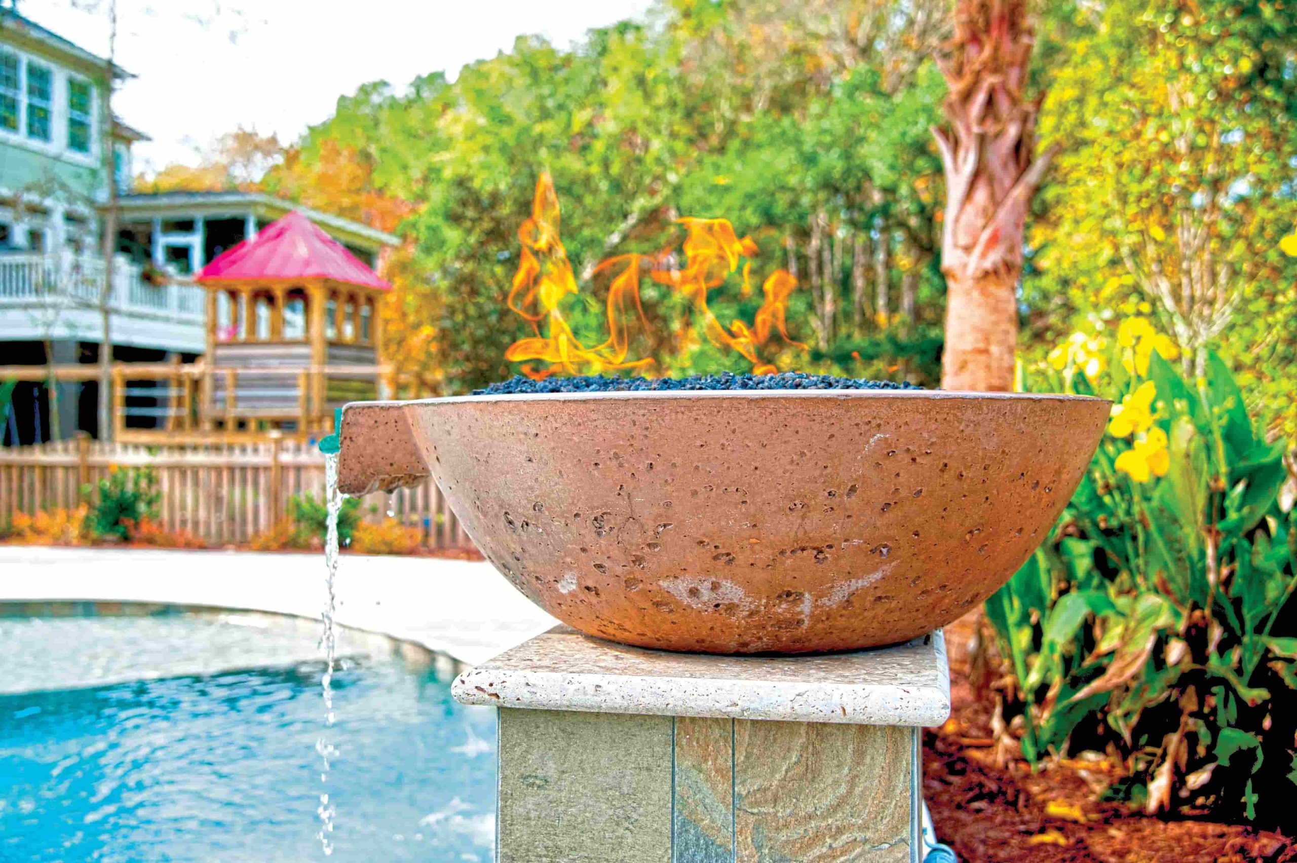 Close-up of a stone fire bowl with flames and a small water spout beside a backyard swimming pool.