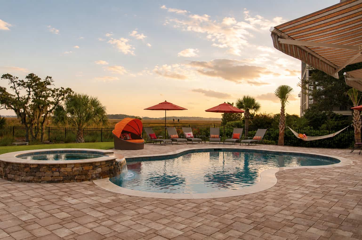 Custom Bluehaven backyard swimming pool and raised stone spa at sunset, with paver patio, loungers, and umbrellas