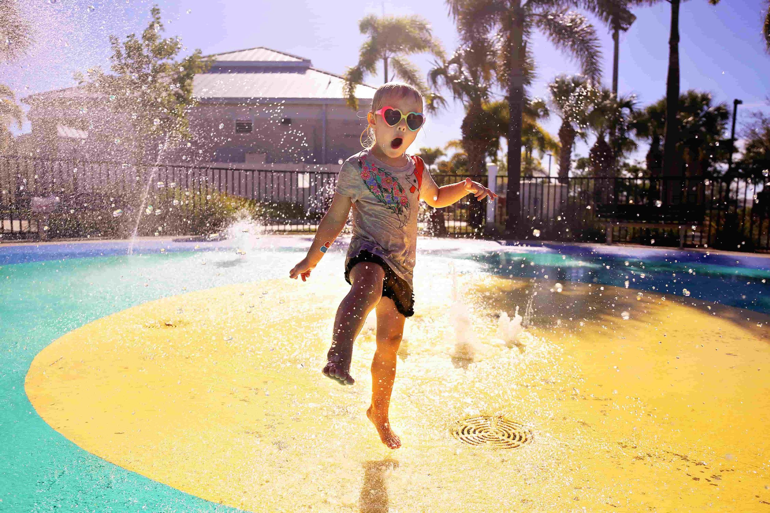 Young child in heart-shaped sunglasses joyfully running through water sprays at a colorful splash pad, with palm trees and sunlit sky in the background.