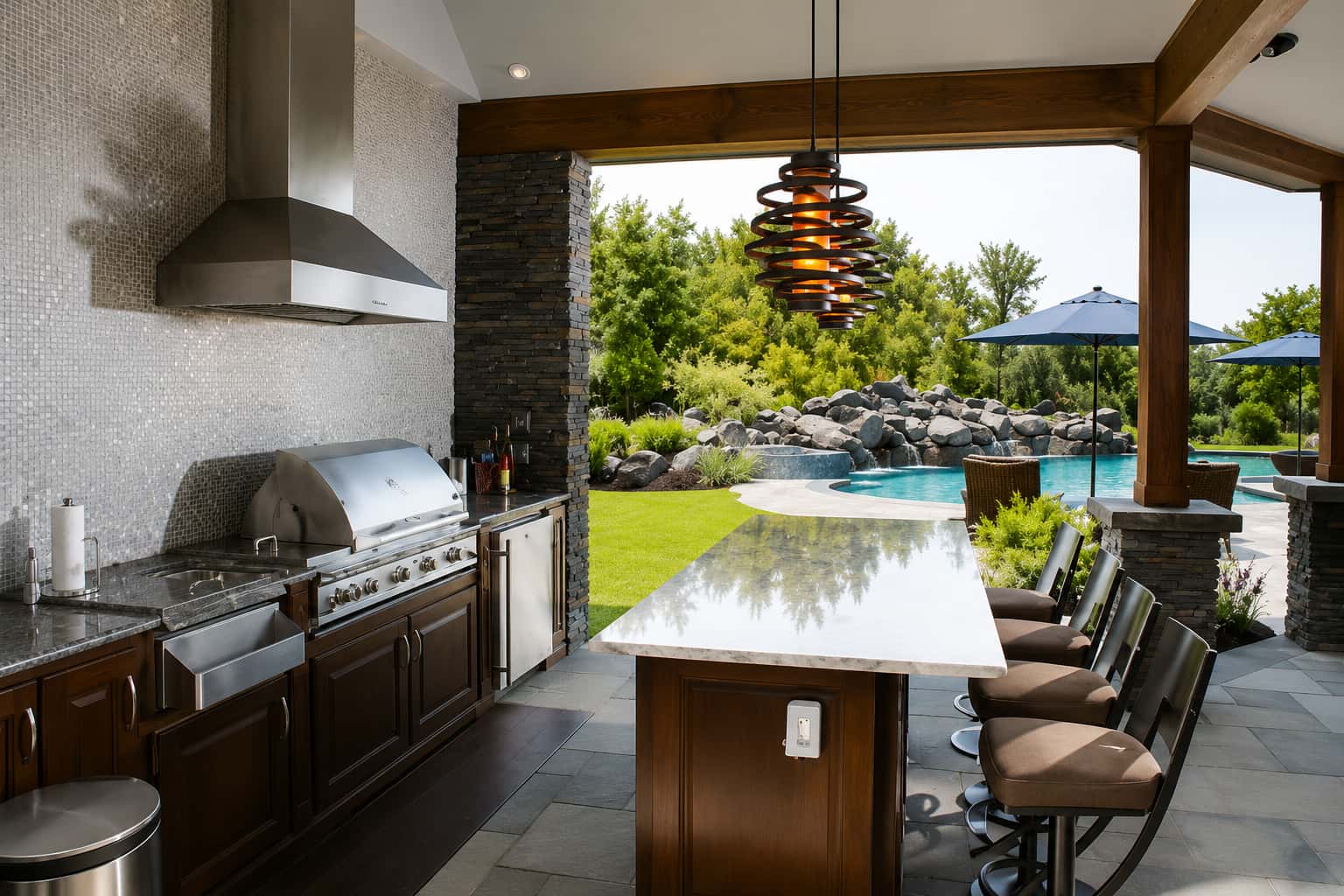Blue Haven covered outdoor kitchen with stainless steel grill, range hood, marble bar with swivel stools, and pendant lighting overlooking a pool with rock waterfall.