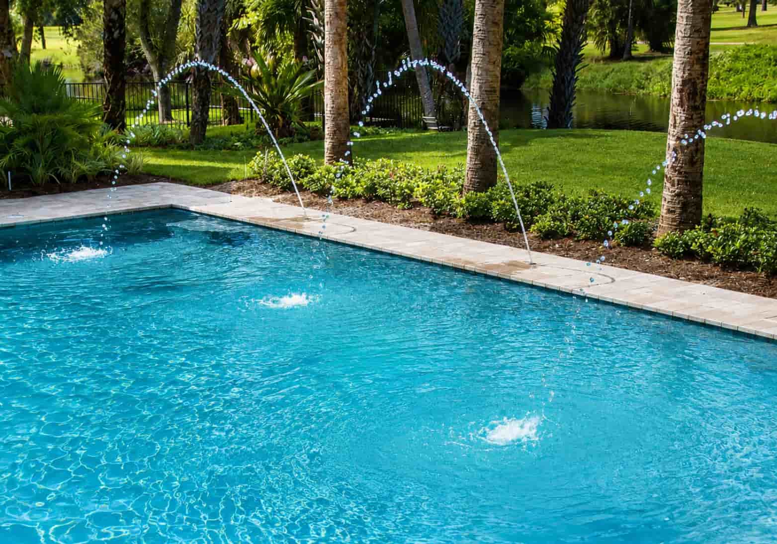 Deck jets shooting arcing water streams over a rectangular pool with tropical palm tree landscaping