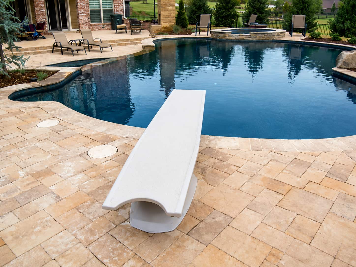 White diving board on a travertine pool deck next to a Blue Haven inground pool and spa