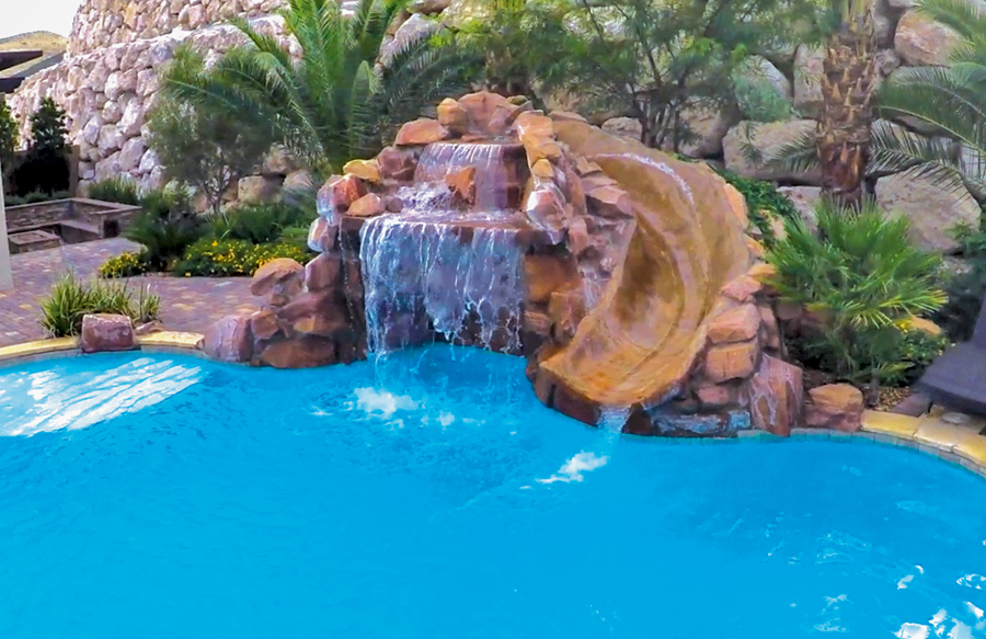 Backyard swimming pool with faux rock waterfall and curved stone slide surrounded by palm trees