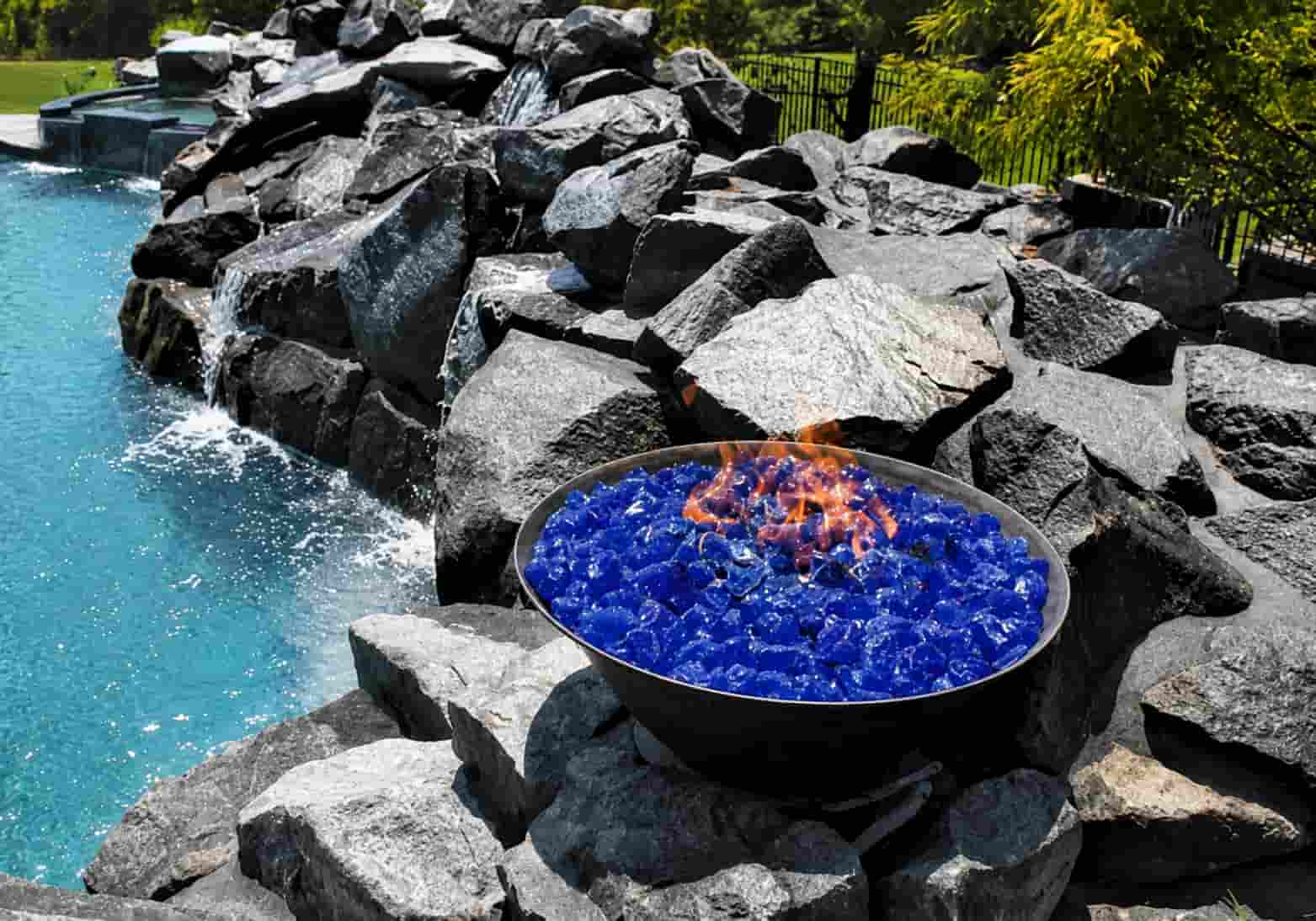 fire bowl filled with blue iridescent fire glass set into a large natural rock waterfall beside a pool
