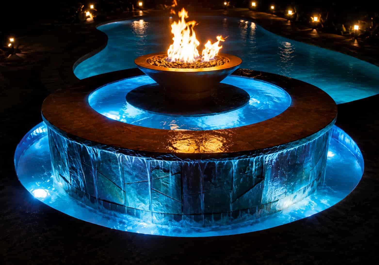 lagoon-style pool at night with two fire bowls on boulder pedestals flanking an illuminated spa and rock waterfall