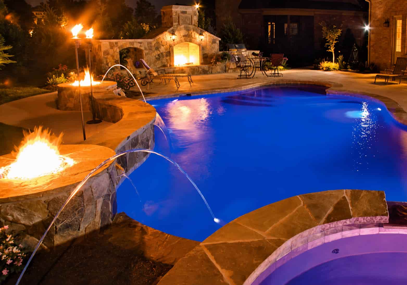 freeform pool at night with a decorative fire pit, arc water jets, a stone outdoor fireplace, and an illuminated spa