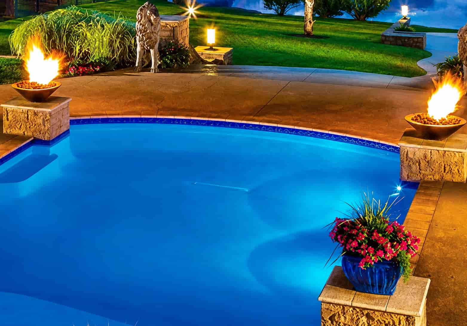 freeform backyard pool at night with two copper fire bowls on stone pedestals and landscape lighting