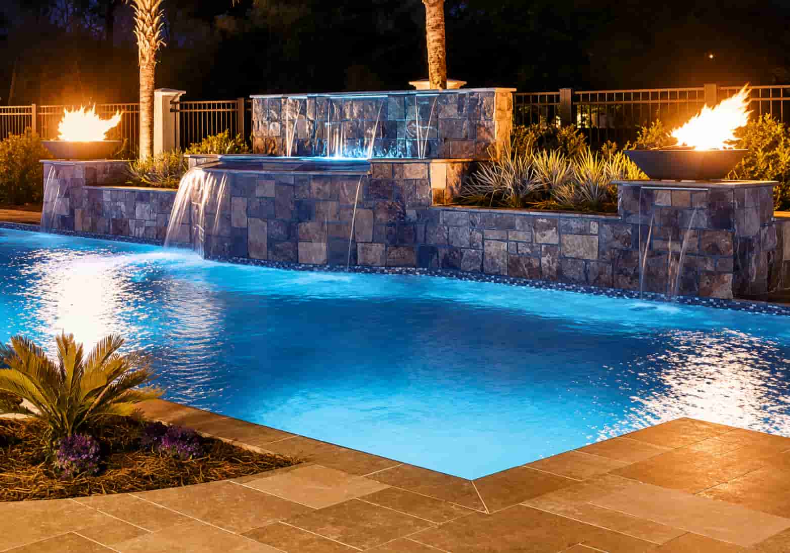 geometric pool at night with a stone waterfall wall flanked by two fire bowls on pillars and blue LED pool lighting