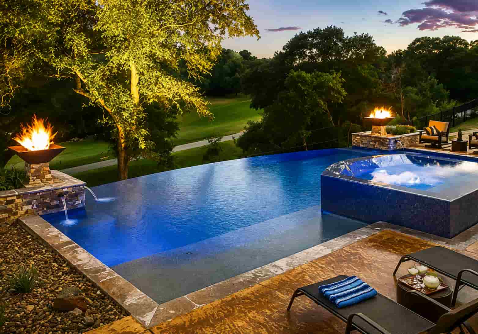 infinity pool at dusk with a copper fire bowl on the left and a stone fire pit near poolside seating on the right