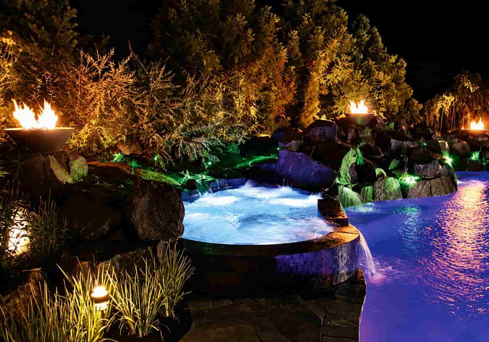 lagoon-style pool at night with fire bowls on boulders, a rock waterfall, illuminated spa, and LED landscape lighting