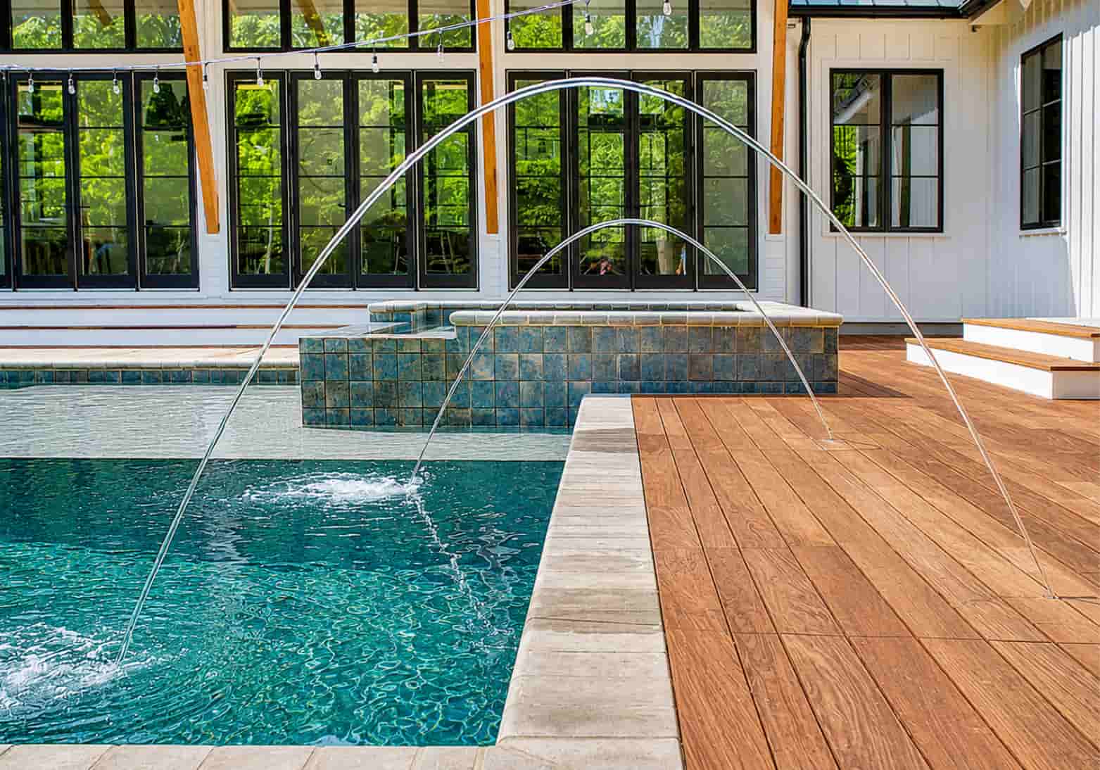 Laminars arcing over a modern farmhouse pool with an elevated tiled spa and wood deck