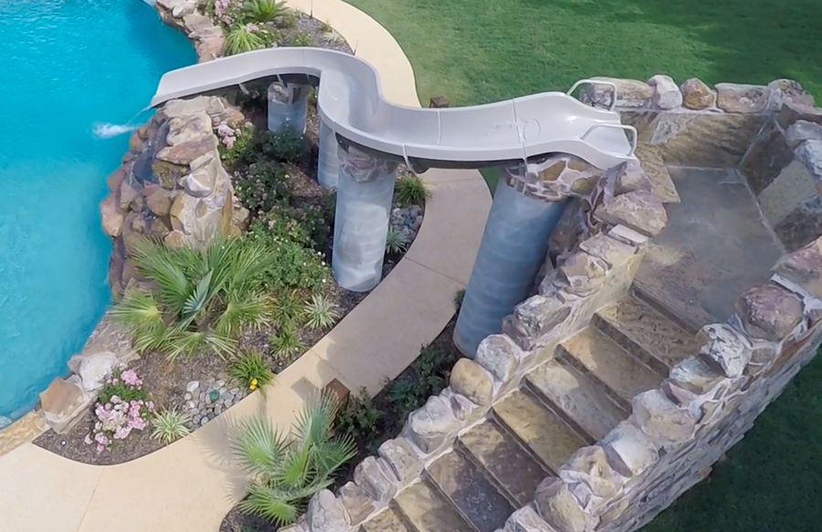 Elevated pool slide supported by columns with stone steps leading up beside landscaped pool