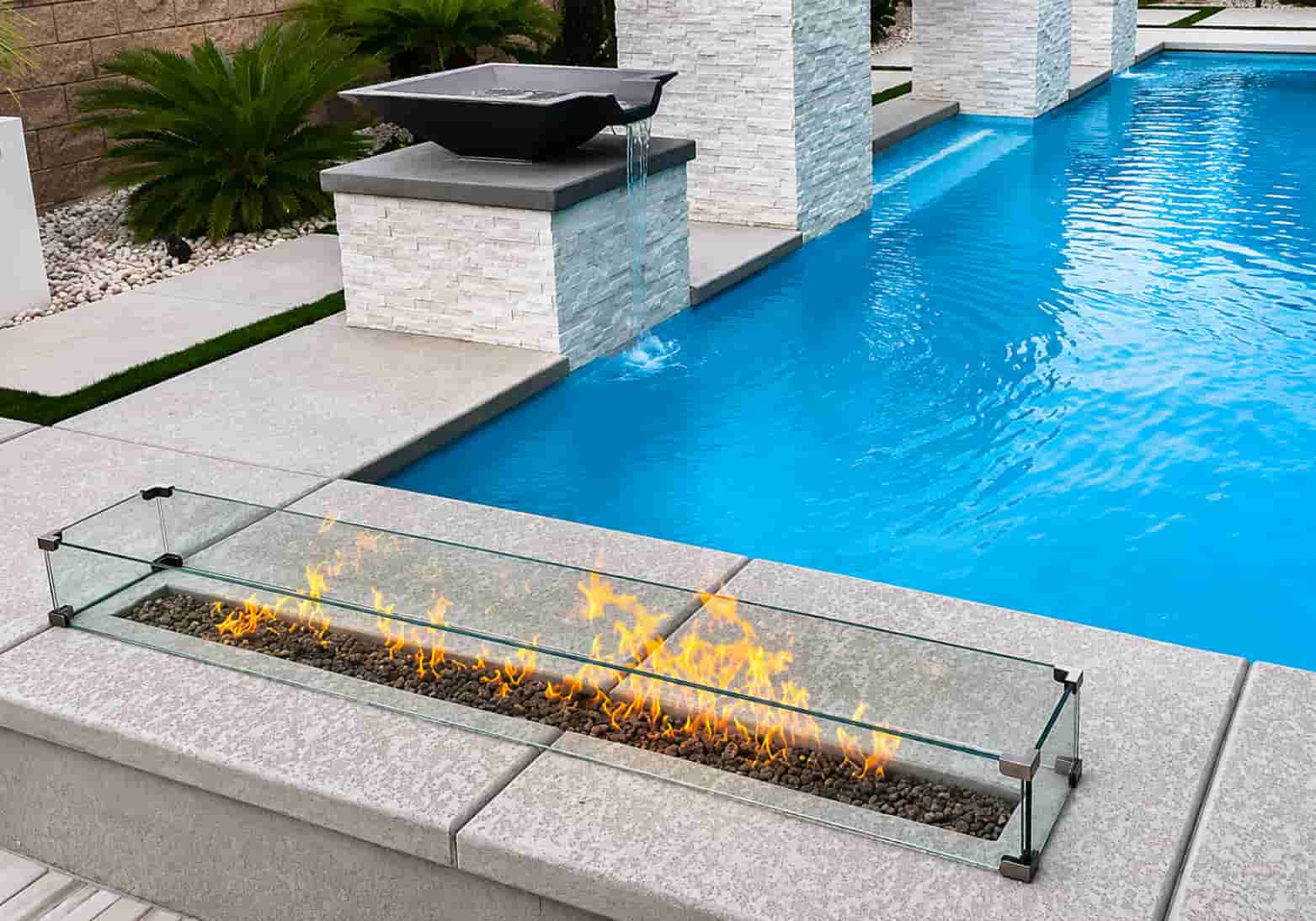 glass-enclosed linear fire box with lava rocks on the deck of a modern backyard pool