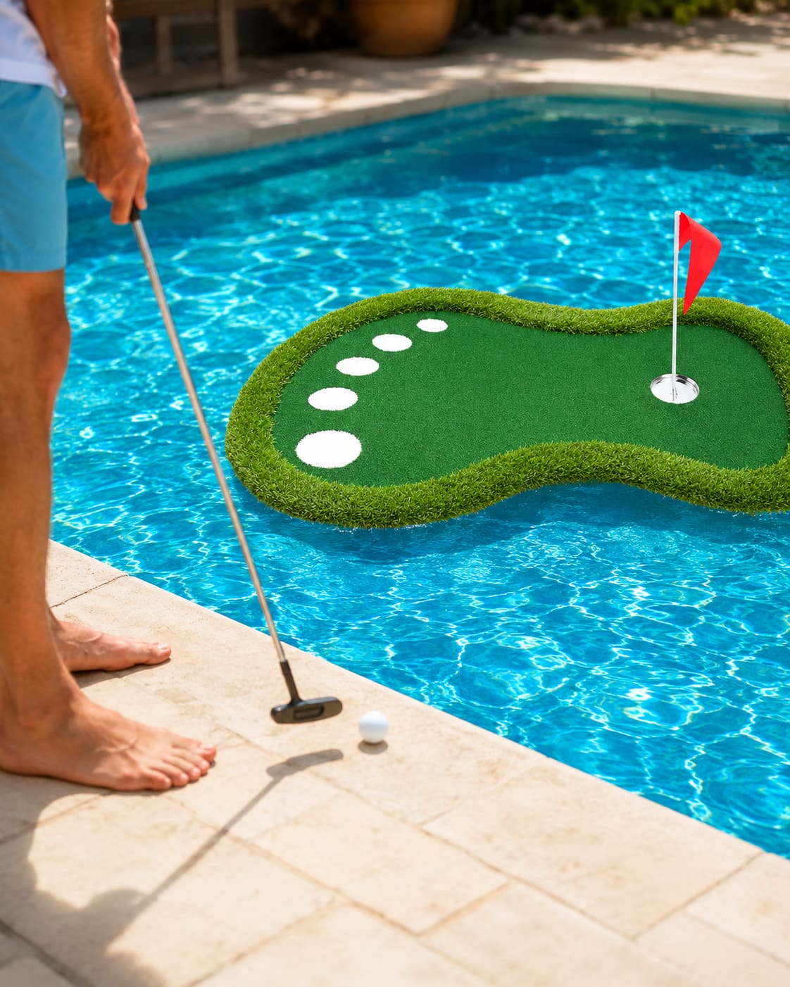 Man on the pool deck chipping a golf ball onto a floating putting green in a backyard pool.