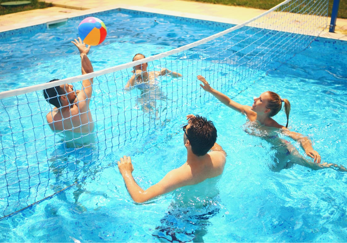 Four friends playing pool volleyball with a beach ball over a net in a backyard swimming pool.