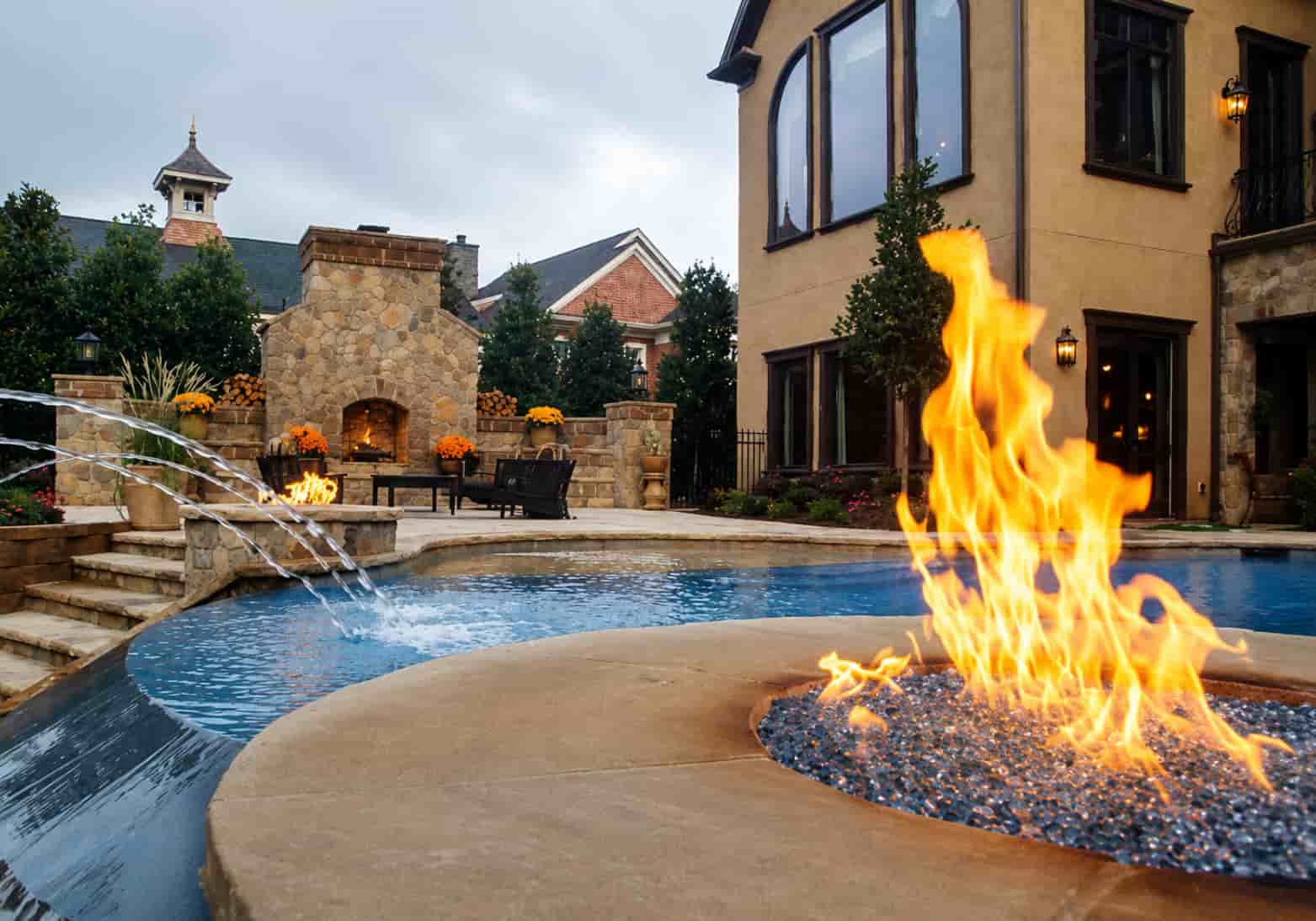 close-up of a pool fire bowl filled with black fire glass beside a pool with water jets and a stone outdoor fireplace