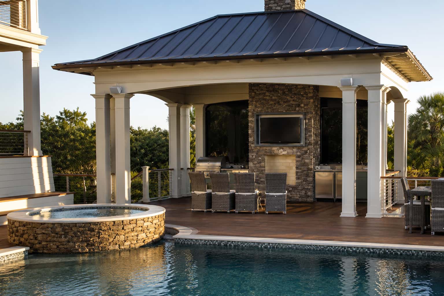 Coastal-style Blue Haven poolside pavilion with white columns, metal roof, stone fireplace, mounted TV, and bar seating beside a pool with raised stone spa.