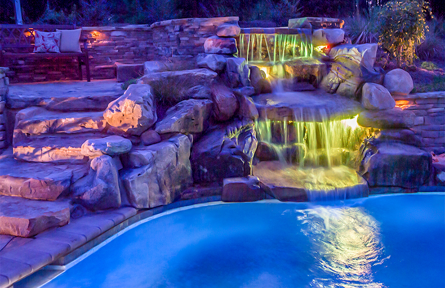 Multi-tier rock waterfall with colored LED lighting on a custom pool at night