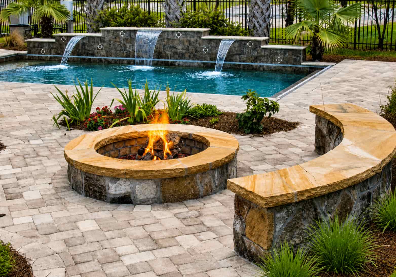 Round fire pit with matching curved stone sitting wall beside pool with waterfalls
