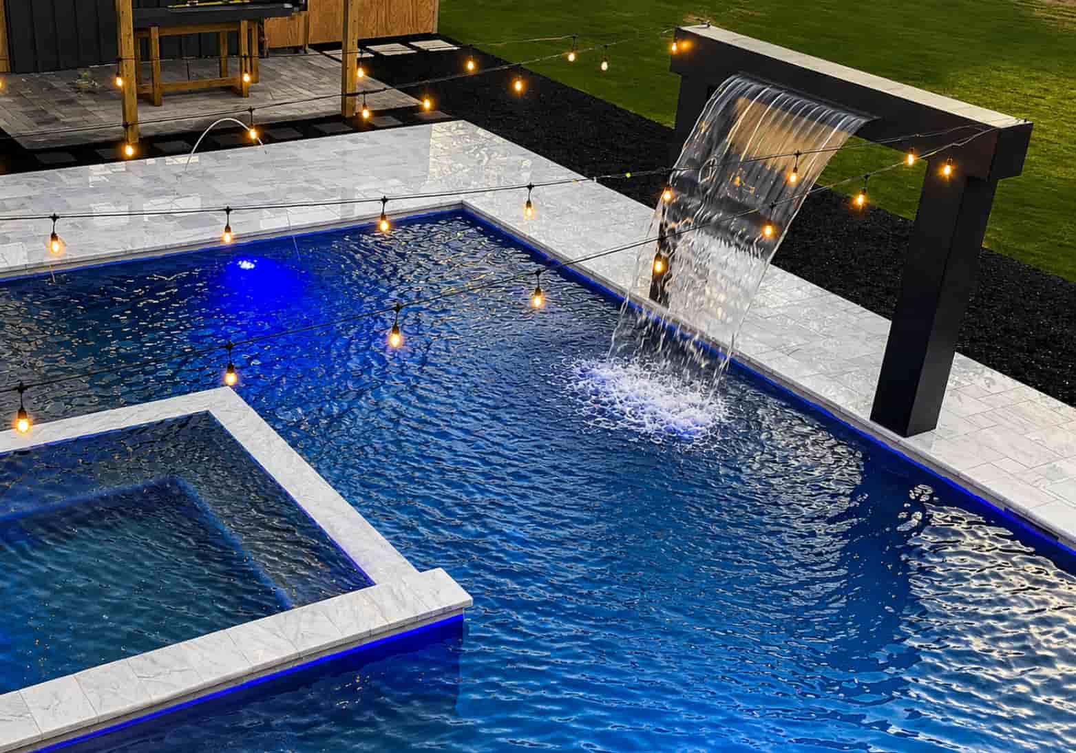 Sheer descent cascade on a black metal frame over a modern pool with white marble tile at night