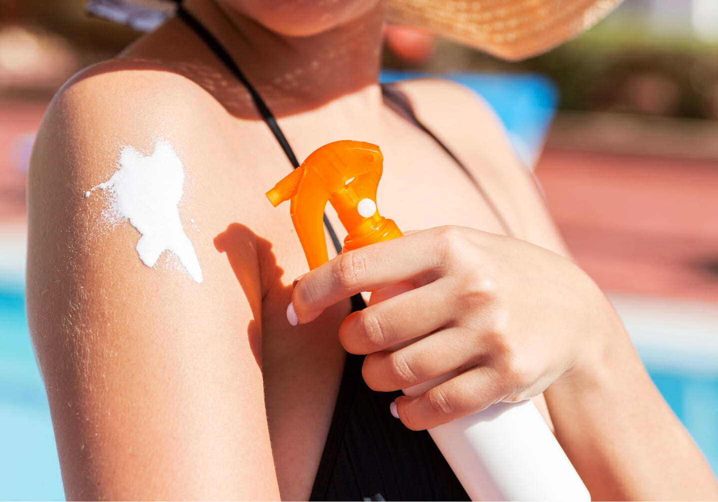 Woman in a sun hat applying spray sunscreen at the pool, leaving an uneven white patch on her shoulder