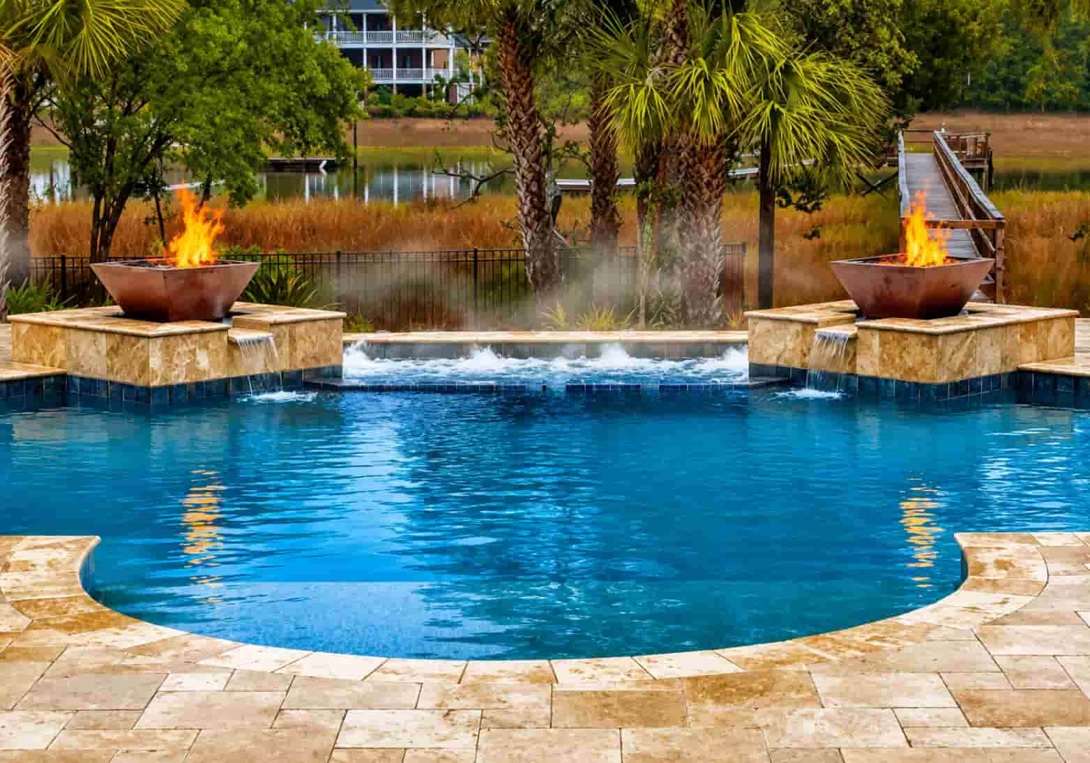 two square copper fire bowls on travertine pedestals flanking a freeform pool with spa spillover and waterfront background
