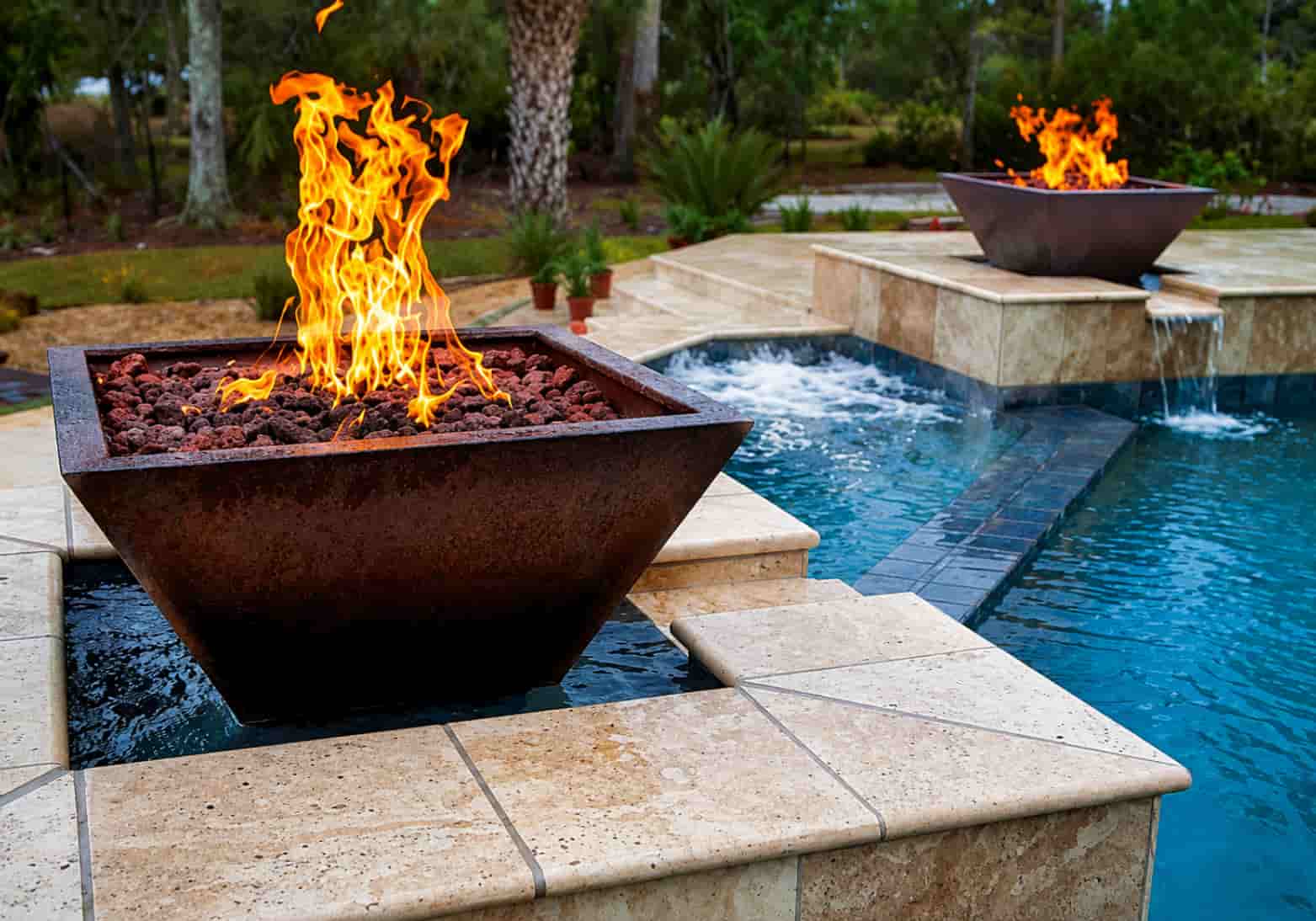 two square copper fire bowls with lava rocks on travertine pedestals beside a rectangular pool with sheer descent water features