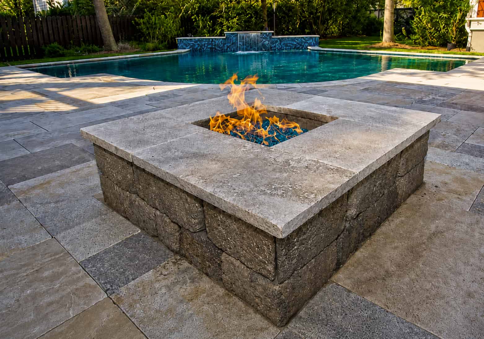 Square fire pit with travertine cap and blue fire glass on matching travertine pool deck