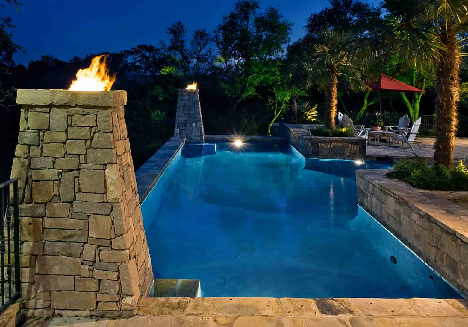 two stone pillar fire features flanking a geometric infinity-edge pool at night with palm trees and landscape lighting