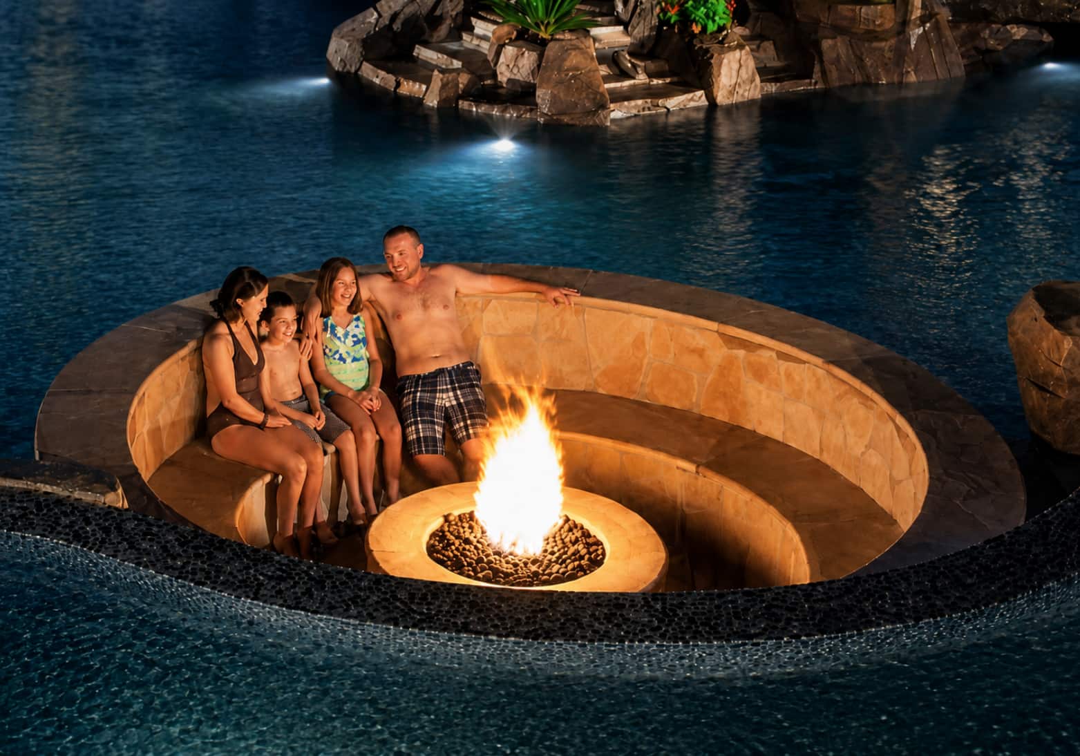 Family sitting in sunken conversation pit with fire built into lagoon swimming pool at night