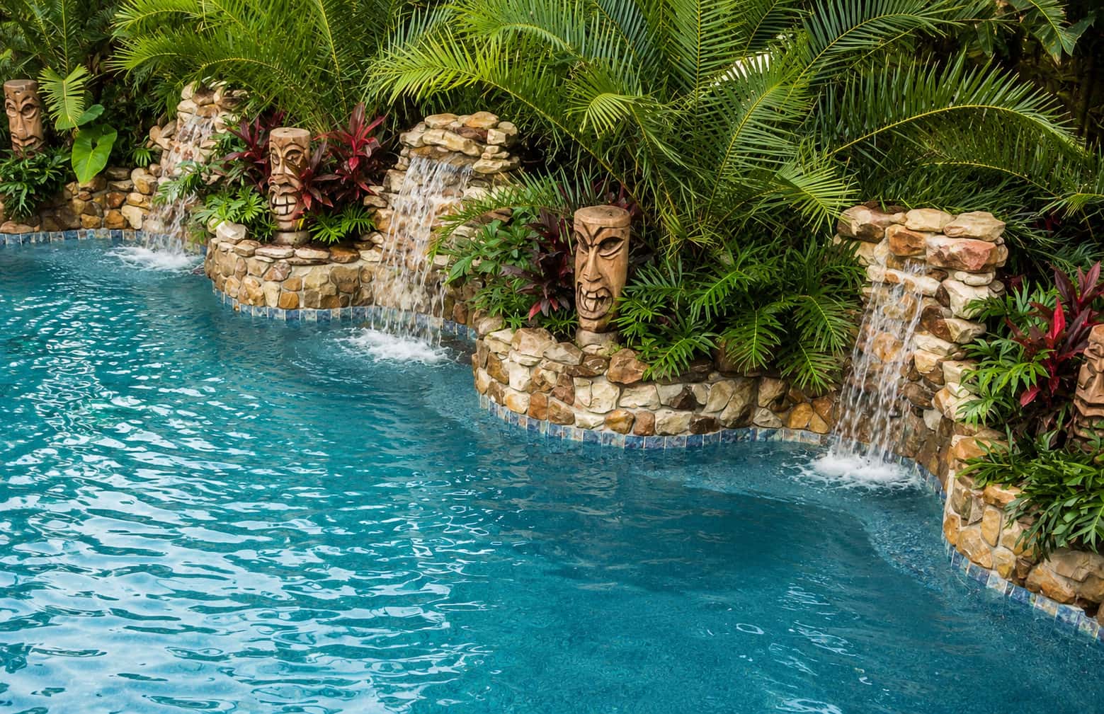 Tropical backyard pool with turquoise water, stone waterfalls, tiki-style statues, and dense green landscaping.