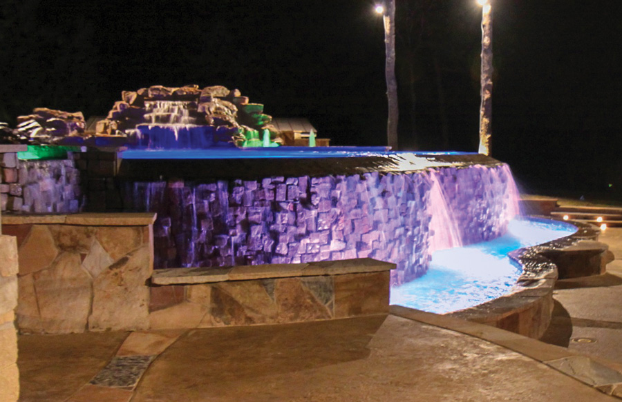 Infinity edge pool with violet LED lighting on stone dam wall and blue-lit overflow catch basin at night